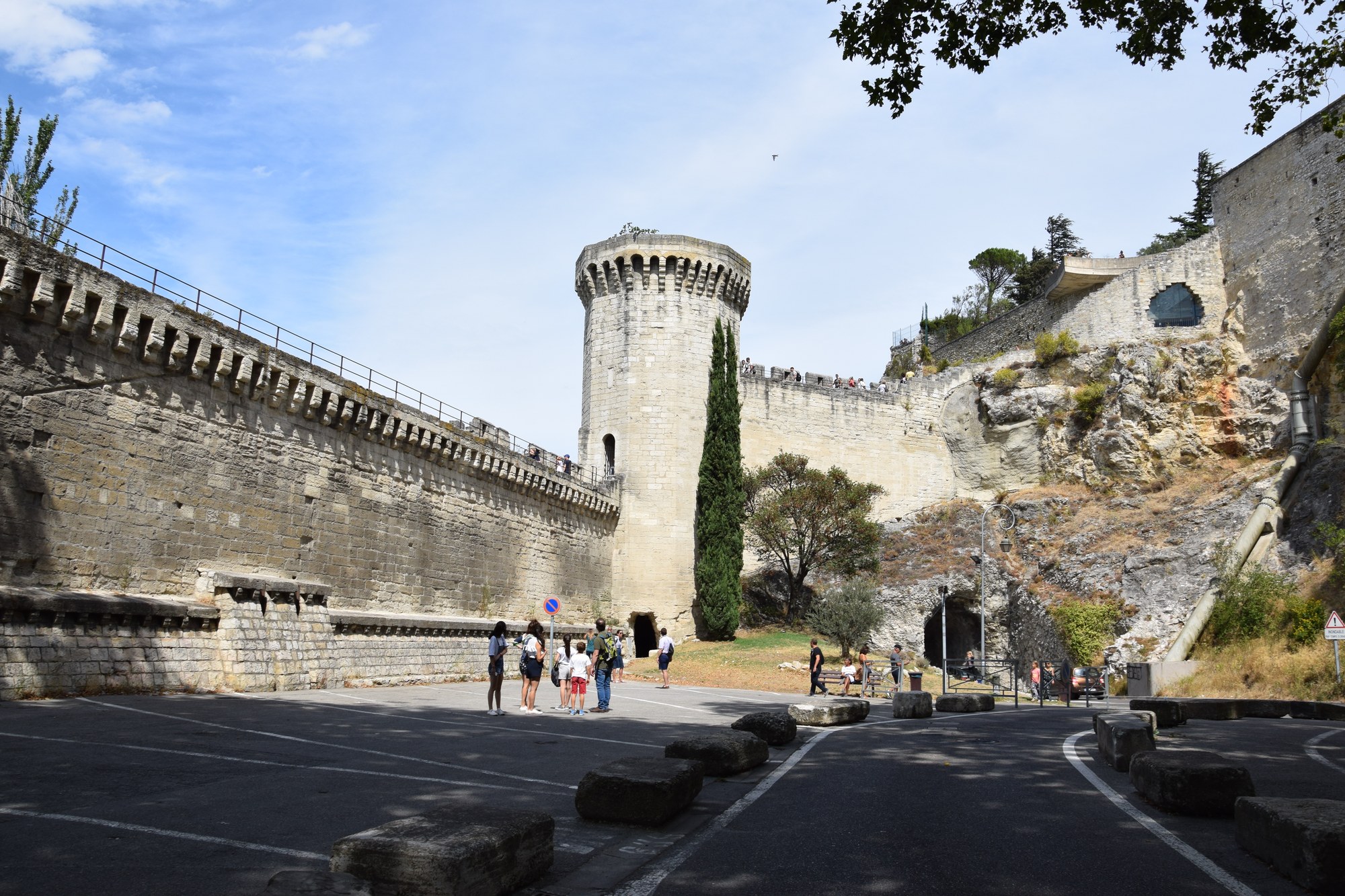 Les remparts d'Avignon et leur tour ronde un après-midi d'août, herbe jaunie au pied du rocher, cyprès et oliviers, passants à l'ombre