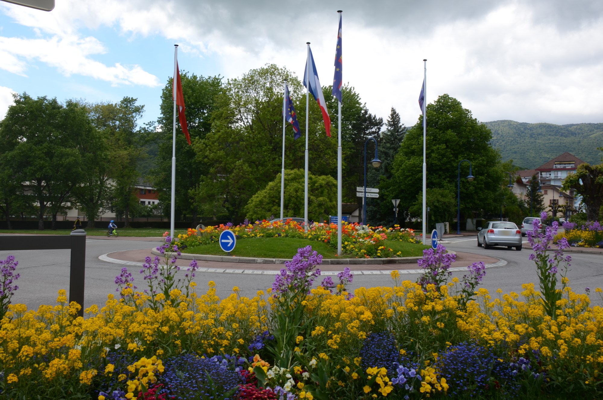 Un rond-point fleuri de Divonne-les-Bains où flottent côte à côte les drapeaux suisse, français et européen, montagnes du Jura en arrière-plan