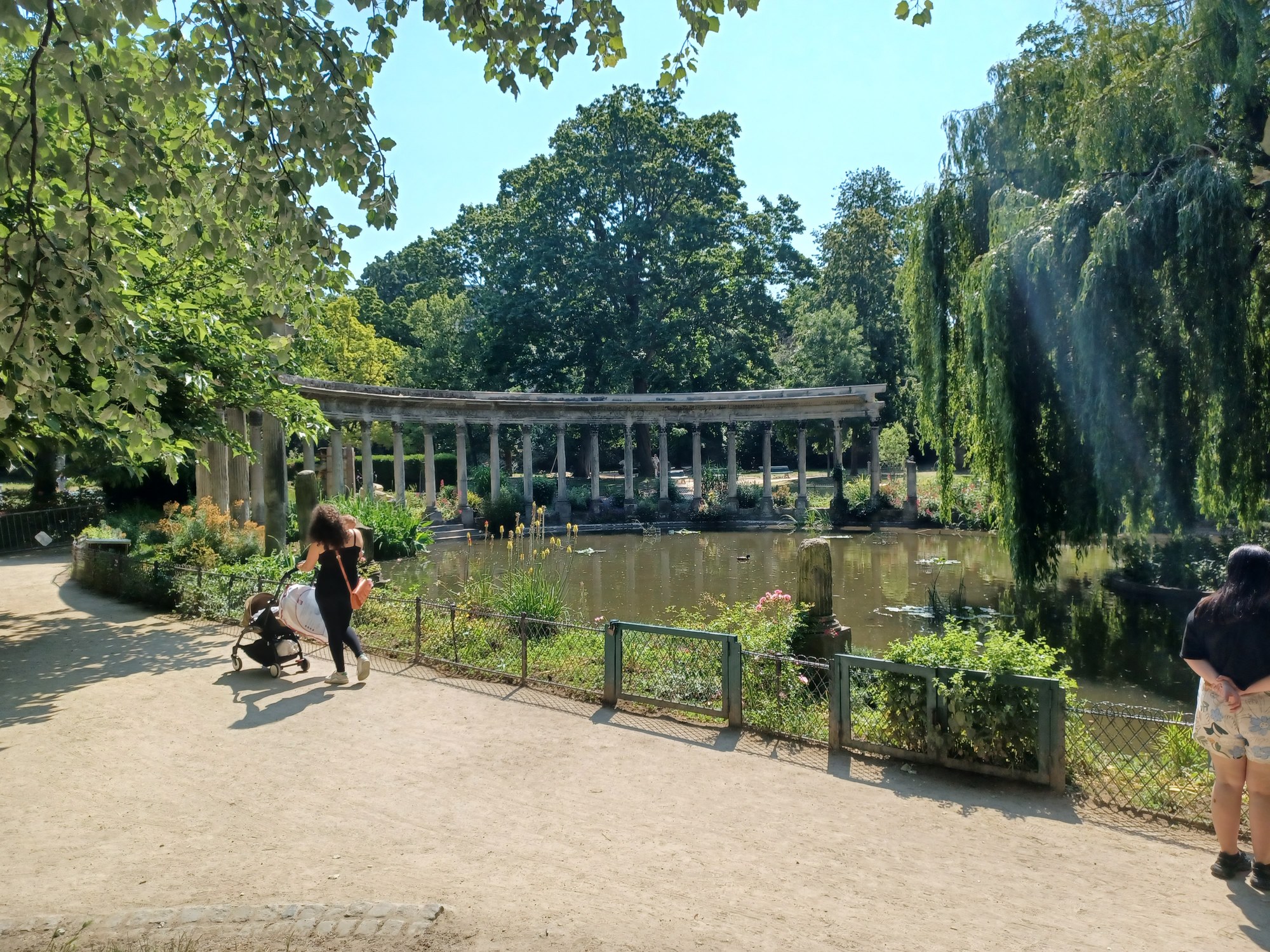 La colonnade du parc Monceau au bord du bassin, un jour d'été : promeneurs sur l'allée, saule et verdure sous un ciel bleu