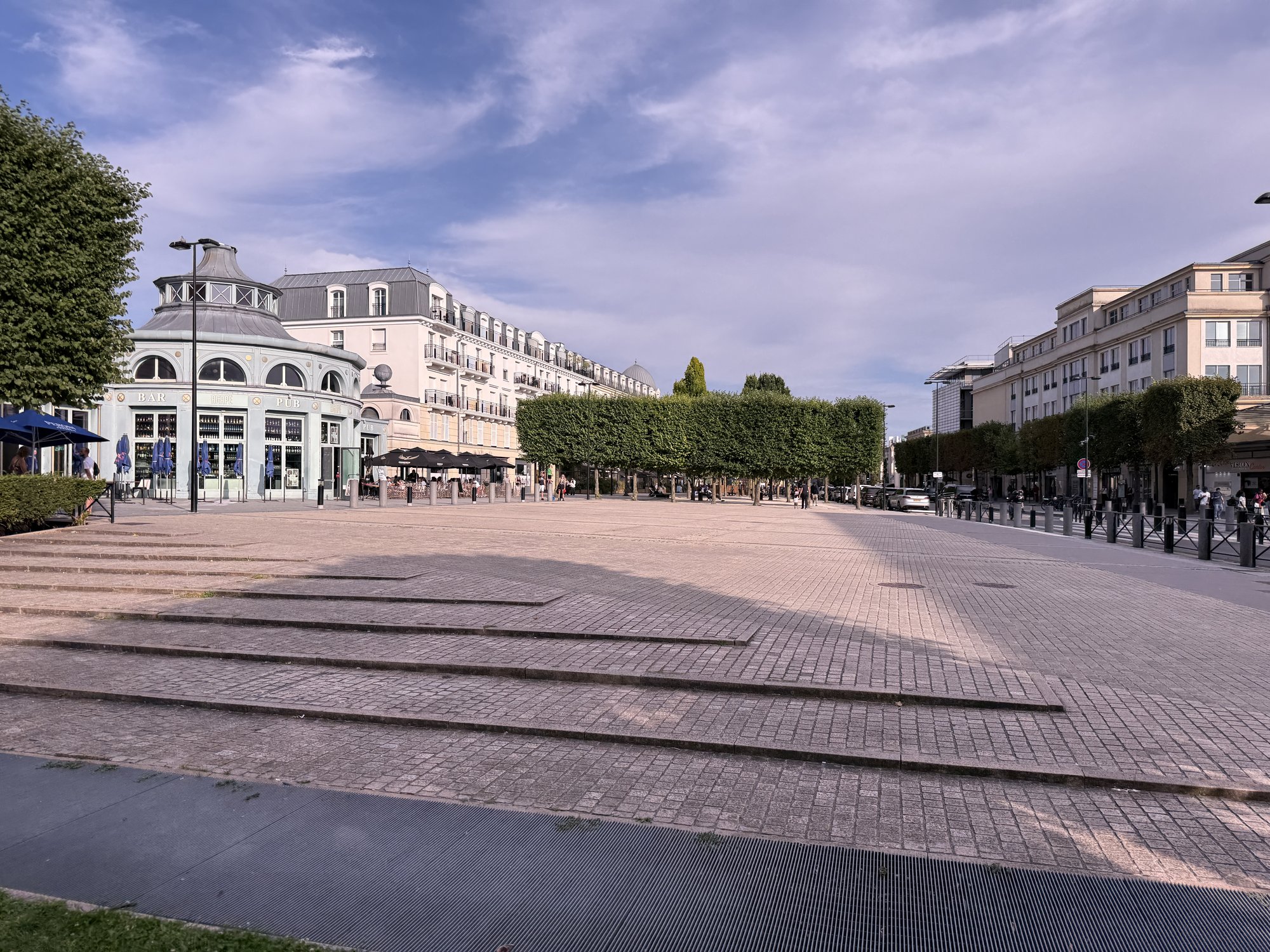 La place d'Ariane à Serris : parvis pavé, arbres taillés, immeubles de logements récents et terrasses de café
