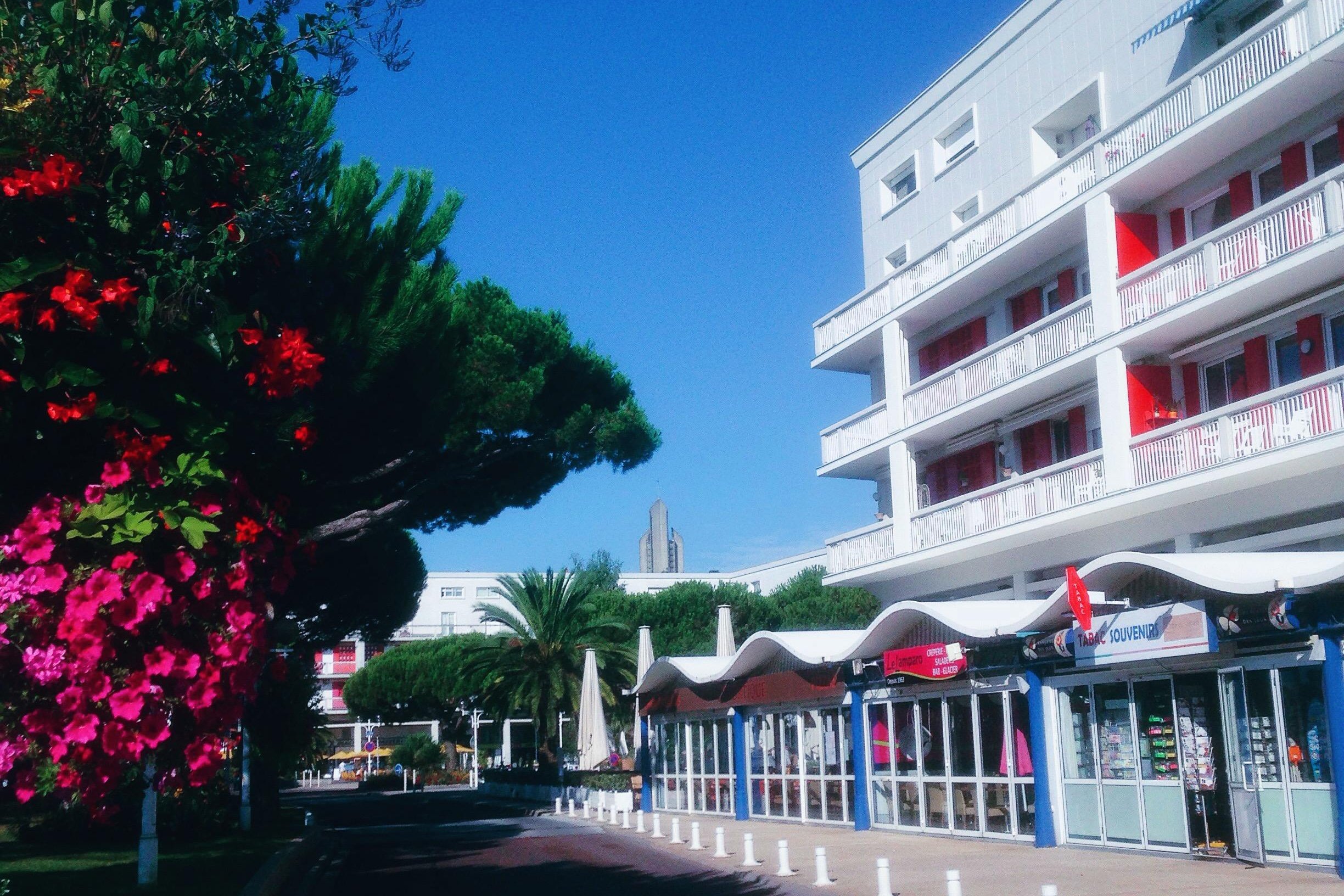 Le front de mer de Royan : immeuble blanc de la Reconstruction aux balcons rouges, commerces sous auvents et arbres en fleurs