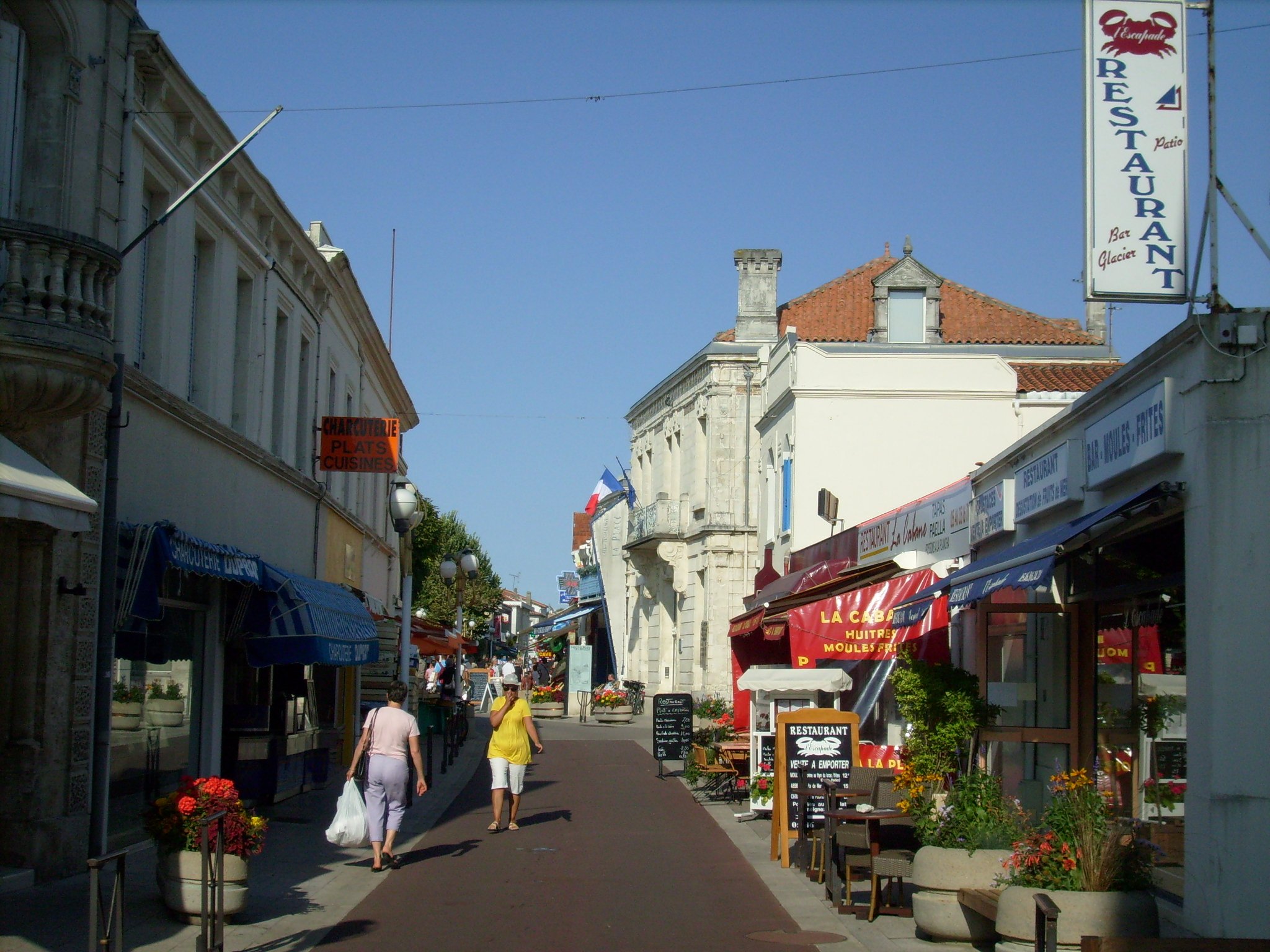 Une rue commerçante piétonne du centre de Saint-Georges-de-Didonne : boutiques, terrasses et jardinières fleuries sous le ciel d'été