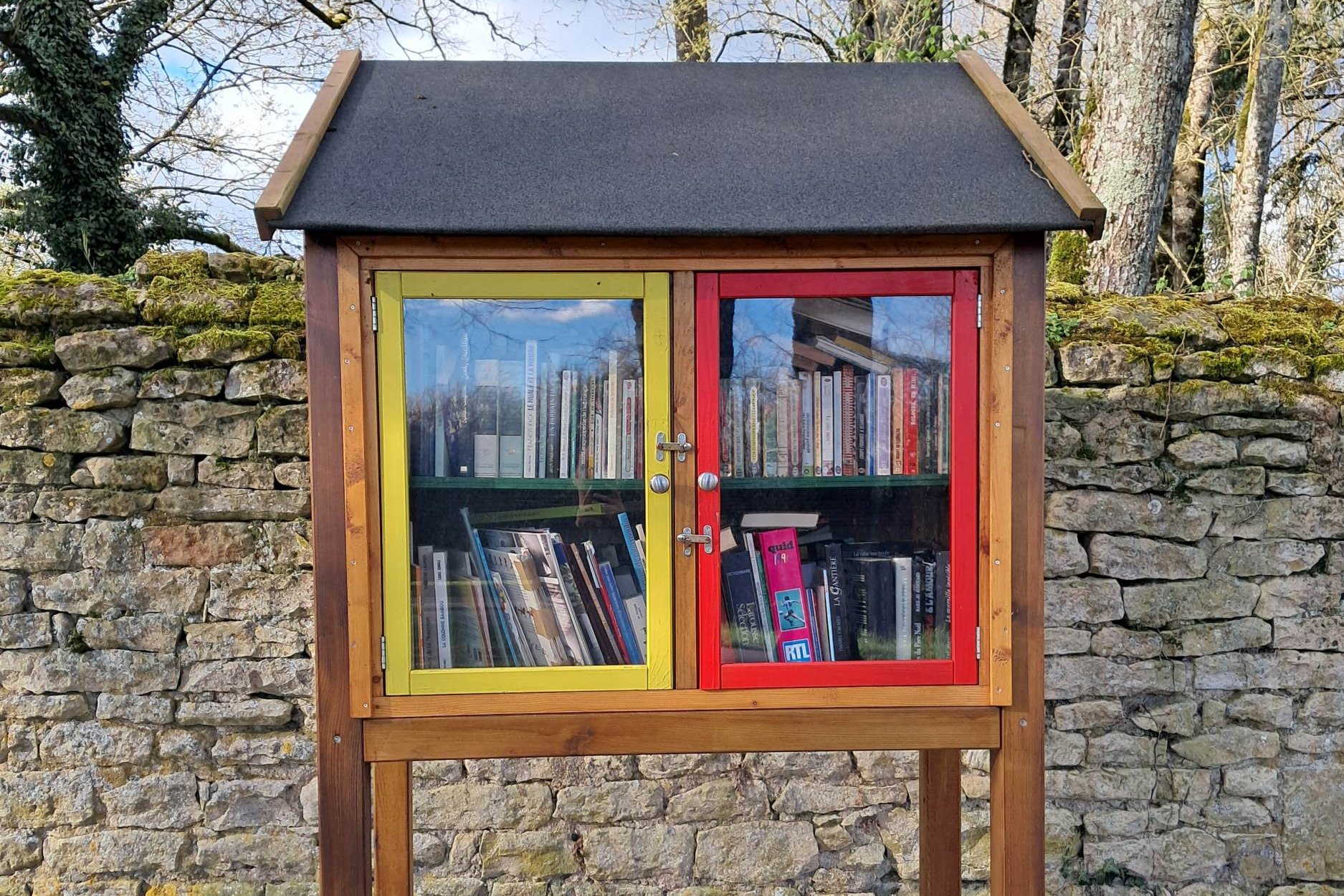 La boîte à livres de la rue de l'Église à Urzy, adossée à un mur de pierres, sous les arbres